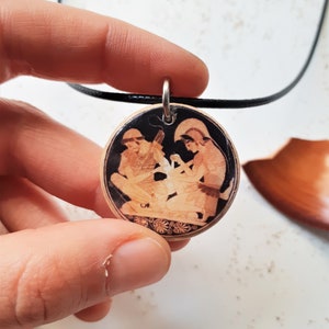 May include: A round pendant necklace with a black cord. The pendant features a black and white image of two figures in a scene from Greek mythology. The pendant is made of wood and has a detailed design.
