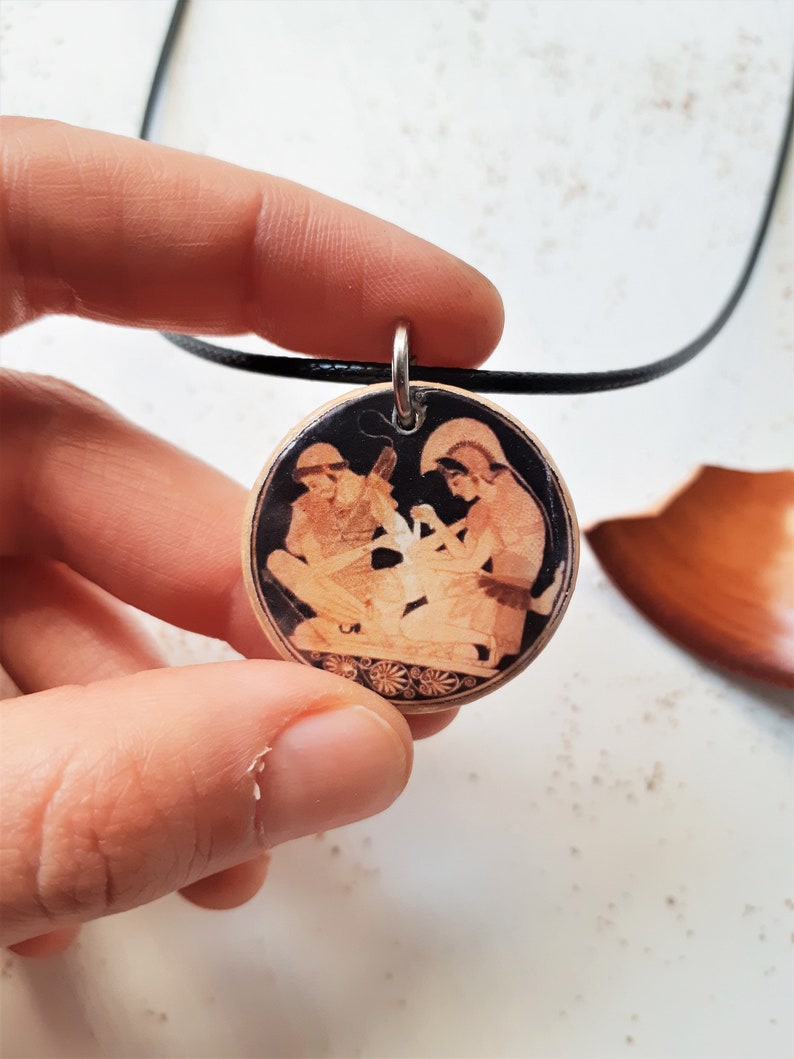 May include: A round pendant necklace with a black cord. The pendant features a black and white image of two figures in a scene from Greek mythology. The pendant is made of wood and has a detailed design.