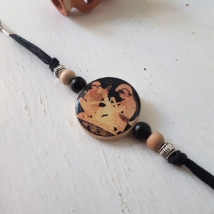 Puede incluir: Un brazalete de madera negro y marr&oacute;n con un colgante redondo que presenta una imagen en blanco y negro de dos figuras. El colgante est&aacute; rodeado de cuentas negras y marrones.