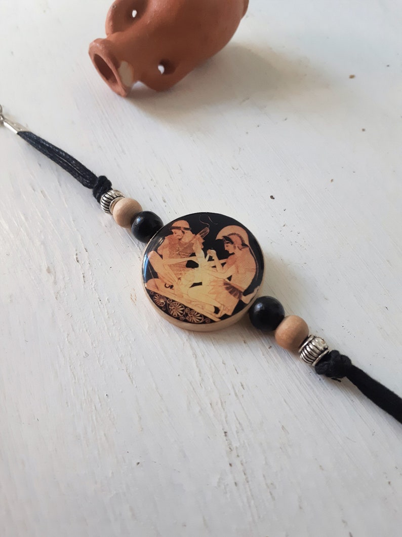 Puede incluir: Un brazalete de madera negro y marr&oacute;n con un colgante redondo que presenta una imagen en blanco y negro de dos figuras. El colgante est&aacute; rodeado de cuentas negras y marrones.