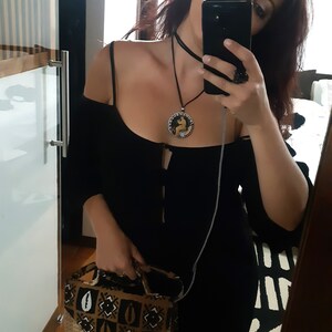 Peut inclure: Robe noire &agrave; &eacute;paules d&eacute;nud&eacute;es, ras du cou et collier pendentif assortis. Le pendentif a un motif circulaire avec une figure &agrave; l'int&eacute;rieur. Un sac &agrave; main &agrave; motifs avec une base tiss&eacute;e est tenu &agrave; la main.