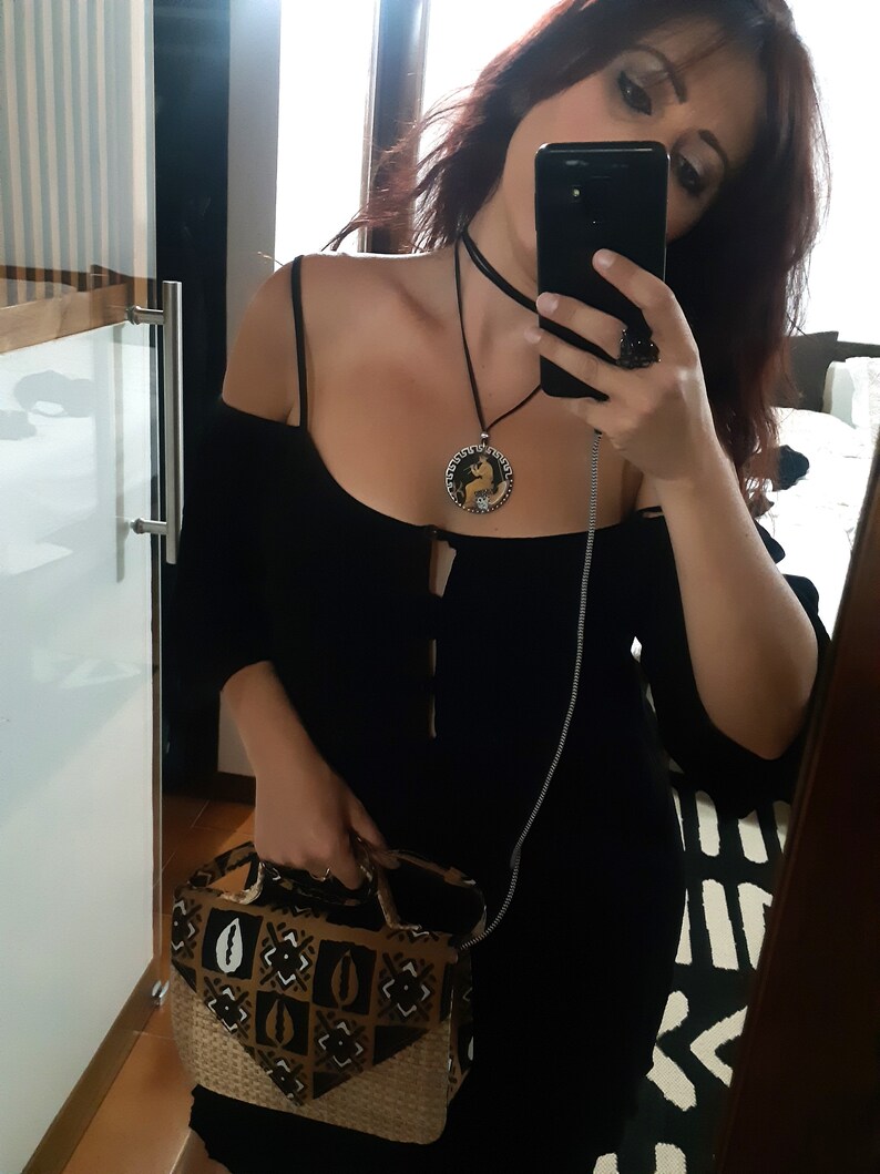 Peut inclure: Robe noire &agrave; &eacute;paules d&eacute;nud&eacute;es, ras du cou et collier pendentif assortis. Le pendentif a un motif circulaire avec une figure &agrave; l'int&eacute;rieur. Un sac &agrave; main &agrave; motifs avec une base tiss&eacute;e est tenu &agrave; la main.