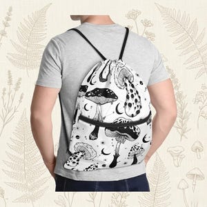 May include: A white drawstring backpack featuring a black and white mushroom design. The bag has a black zipper and black drawstrings. The mushrooms are illustrated with intricate details, and the design includes crescent moons and stars.