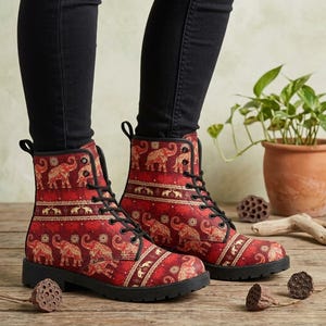 May include: Red and gold elephant print combat boots with black laces and soles. The boots feature a repeating pattern of elephants and decorative borders. The boots are worn with black pants.