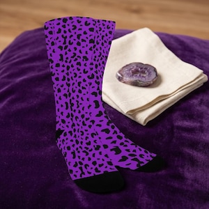 May include: A pair of knee-high socks with a purple background and black leopard print pattern. The socks have black heels and toes. A folded beige cloth and a purple geode are in the background.