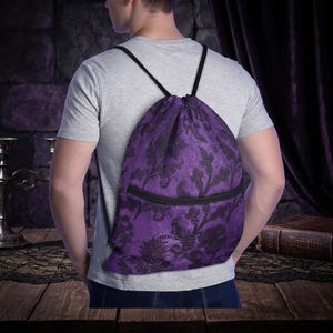 May include: A purple drawstring bag featuring a black floral design and a black zipped pocket. The bag has black straps and is made of textured fabric. The backpack is being worn by a person.