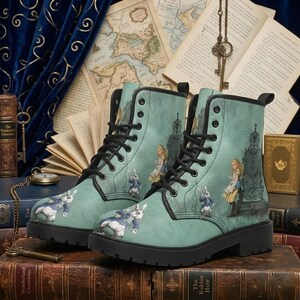 Könnte beinhalten: Ein Paar tealfarbene Kampfstiefel mit Illustrationen aus Alice im Wunderland. Die Stiefel haben schwarze Schnürsenkel und Sohlen, mit Bildern von Alice, dem weißen Kaninchen und einem Thron. Die Stiefel stehen vor einem Hintergrund aus Büchern und Vintage-Karten.