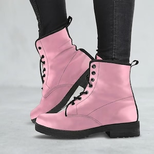 May include: Pink combat boots with black laces and soles. The boots have a smooth, glossy finish and a classic lace-up design. The boots are worn with black jeans.
