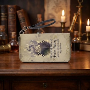 May include: A beige clutch wallet with the text "Encyclopedia of Potion Making" and a cauldron illustration. It has a black wrist strap and a zip closure. The design has a vintage, aged appearance, reminiscent of an old book.