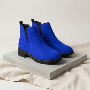 Könnte beinhalten: Ein Paar leuchtend blaue Stiefeletten mit Wildleder-Optik. Die Stiefel haben einen niedrigen, schwarzen Absatz und einen V-förmigen Ausschnitt an der Seite. Sie stehen auf einem grauen Betonblock vor neutralem Hintergrund. Die Stiefel sind ca. 15 cm hoch.