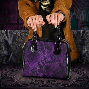 May include: A purple handbag with a floral design and black detailing. The bag features a structured shape, a top handle, and a shoulder strap. The bag is held by a person wearing a brown jacket.