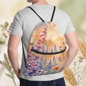 Könnte beinhalten: Ein Turnbeutel mit Kordelzug, der ein Aquarelldesign eines Wildblumenfeldes in Rosa-, Lila- und Orangetönen zeigt. Die Tasche hat schwarze Kordelzüge und eine schwarze Reißverschlusstasche. Der Hintergrund ist ein warmer Sonnenuntergang.