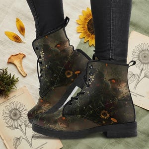 May include: Lace-up combat boots with a dark green and brown floral and mushroom design. The boots have a black sole and heel. The design includes sunflowers and other botanical elements. The boots are worn with black jeans.