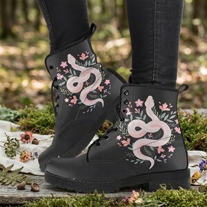 May include: Black combat boots featuring a pink snake and floral design. The boots have a lace-up closure and a sturdy sole. The snake and floral design is printed on the side of the boots. The boots are shown outdoors.