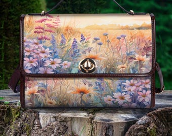 Könnte beinhalten: Eine braune Ledertasche mit einem Blumendruck, der eine Aquarellmalerei eines Blumenfeldes zeigt. Die Tasche hat einen Henkel oben und einen Schultergurt.