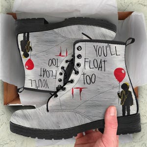 May include: White leather boots with black laces and a black sole. The boots have a graphic print of a clown holding a red balloon and the text "You'll Float Too" on the side.