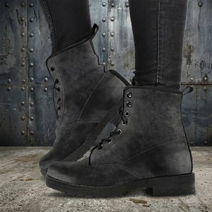 May include: Black combat boots with a textured, distressed appearance. The boots have a lace-up design with metal eyelets and a small heel. The boots are worn with black jeans, creating a monochromatic look.