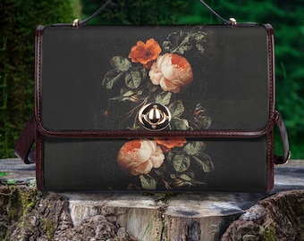 Può includere: Una borsa a tracolla nera con una stampa floreale di rose arancioni e crema e foglie verdi. La borsa ha un bordo marrone, un manico superiore e una chiusura dorata. La borsa è su un ceppo d'albero.
