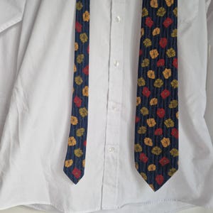 May include: A navy blue tie with a repeating pattern of red, yellow, and orange leaf shapes. The tie is displayed against a white button-down shirt.