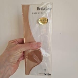 Puede incluir: Un paquete sellado de medias Berkshire micro-stretch en Riviera Tan. El paquete está en plástico transparente y tiene una pegatina dorada con "Riviera Tan" y "8-9". La marca "Berkshire" está impresa en la parte superior.