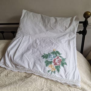 May include: White pillowcase with a floral design featuring pink, blue, and yellow flowers with green leaves. The pillowcase has a crocheted lace trim along the bottom edge. The pillow is on a bed with a decorative headboard.