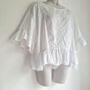 Puede incluir: Una blusa blanca holgada con cuello redondo y mangas cortas con volantes. La blusa presenta detalles bordados y un dobladillo con volantes. La tela parece ligera y aireada, adecuada para climas cálidos. La blusa se muestra en un maniquí.