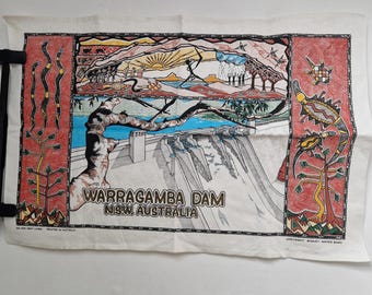 Op de afbeelding: Een witte linnen theedoek met een kleurrijke illustratie van de Warragamba-dam in New South Wales, Australië. De illustratie toont een dam, een rivier, bomen en een zonsondergang. De tekst "Warragamba Dam NSW Australia" is gedrukt in bruine letters.