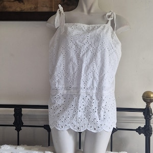 Puede incluir: Top blanco calado con tirantes anudados. El top presenta un escote cuadrado y un dobladillo festoneado. La tela está detallada con calados geométricos y florales. La prenda se exhibe en un maniquí.