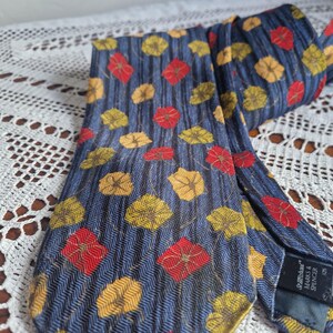 May include: A navy blue tie with a repeating pattern of red and yellow floral designs. The tie has a textured appearance and is folded over on a white lace surface.