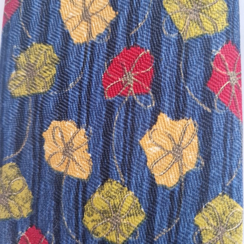 May include: Close-up of a blue fabric with a repeating pattern of stylized leaves in yellow, red, and green. The leaves have a detailed, textured appearance, and are connected by thin, gold-colored lines. The background has a subtle herringbone weave.