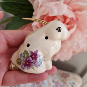 Op de afbeelding: Een crèmekleurig keramieken konijnbeeldje met een goudkleurige lus om op te hangen. Het konijn heeft zwarte ogen en is versierd met paarse en blauwe bloemmotieven. Het beeldje is ongeveer 7,6 cm lang.