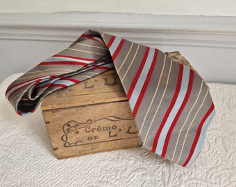 May include: A striped tie in shades of gray, red, light blue, and yellow rests on a wooden box. The tie features a classic pattern of vertical stripes. The box has the text "Crème de L" printed on it.