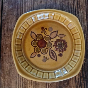 May include: A square, mustard-yellow ceramic plate with a floral design. The plate features a raised, rectangular border and a central design of brown and maroon flowers with leaves. The plate has a vintage aesthetic.