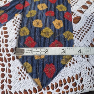 May include: A blue tie with a leaf pattern in red, yellow, and gold. The tie is approximately 3 inches wide. The tie is on a white lace surface.