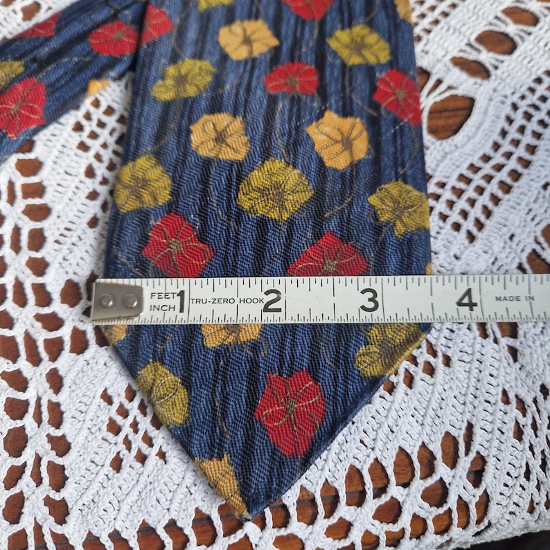 May include: A blue tie with a leaf pattern in red, yellow, and gold. The tie is approximately 3 inches wide. The tie is on a white lace surface.