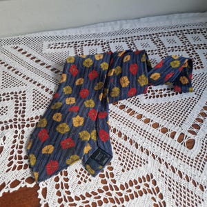 May include: A blue necktie with a repeating pattern of red and yellow leaves. The tie is made of a textured fabric and is displayed on a white lace tablecloth. The tie is a classic accessory.