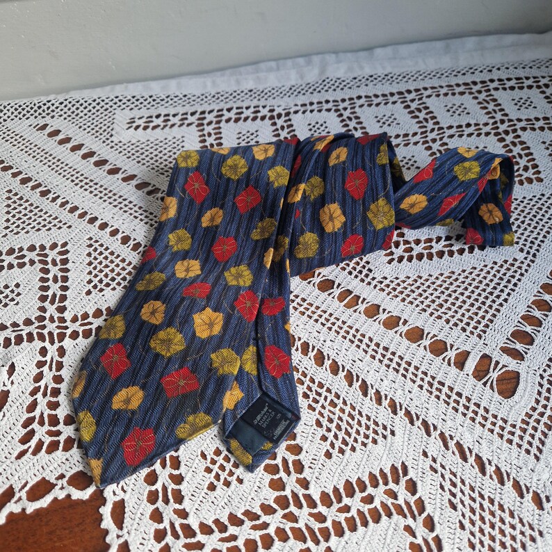 May include: A blue necktie with a repeating pattern of red and yellow leaves. The tie is made of a textured fabric and is displayed on a white lace tablecloth. The tie is a classic accessory.