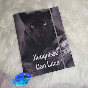 Könnte beinhalten: Ein schwarz-weißes Notizbuch mit einem Pantherbild auf dem Einband. Das Notizbuch trägt den weißen Text "Zeugnisse Can Luca". Ein graues Gummiband sichert das Notizbuch.