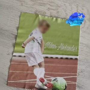 May include: A green and white printed cloth featuring a young soccer player kicking a soccer ball on a track. The name "Milan-Alexander" is printed in cursive. The cloth has a light green border and a logo in the upper right corner.