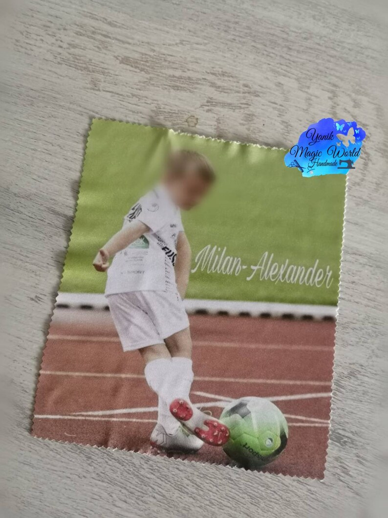 May include: A green and white printed cloth featuring a young soccer player kicking a soccer ball on a track. The name "Milan-Alexander" is printed in cursive. The cloth has a light green border and a logo in the upper right corner.