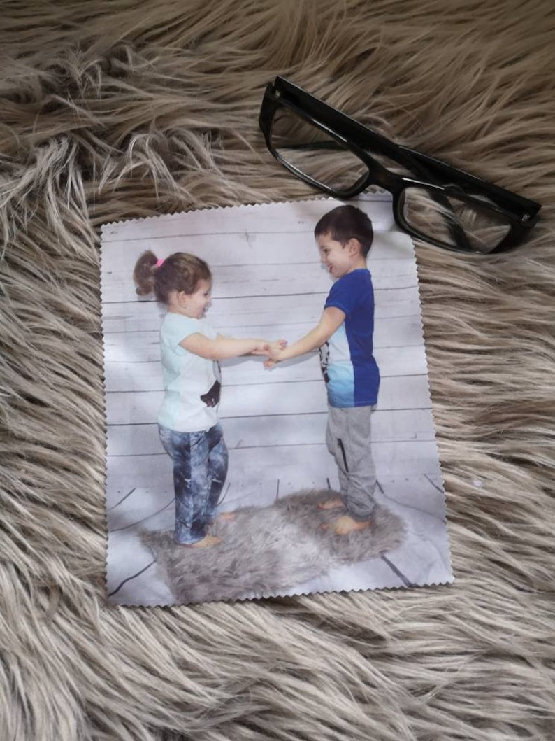 May include: A microfiber cleaning cloth featuring a printed image of two children holding hands. The cloth is rectangular and has a white border. Black-framed eyeglasses rest on the fluffy, light brown surface next to the cloth.