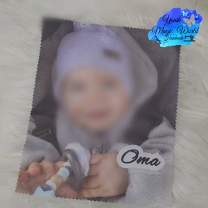 May include: A soft, gray baby blanket with a blurred image of a baby wearing a light blue hat and a gray outfit. The word "Oma" is printed on a white sticker. The blanket has a decorative edge and the "Yanik Magic World Handmade" logo.