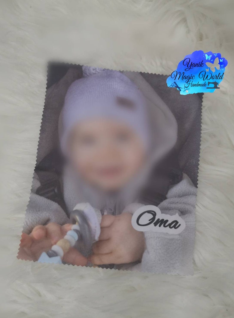 May include: A soft, gray baby blanket with a blurred image of a baby wearing a light blue hat and a gray outfit. The word "Oma" is printed on a white sticker. The blanket has a decorative edge and the "Yanik Magic World Handmade" logo.