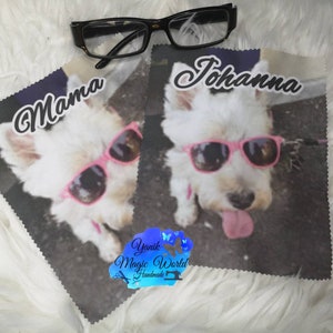 May include: Two microfiber cleaning cloths featuring a white dog wearing pink sunglasses. One cloth has the word "Mama" and the other has the name "Johanna". Black eyeglasses rest on top of the cloths. The cloths are on a white, fluffy surface.