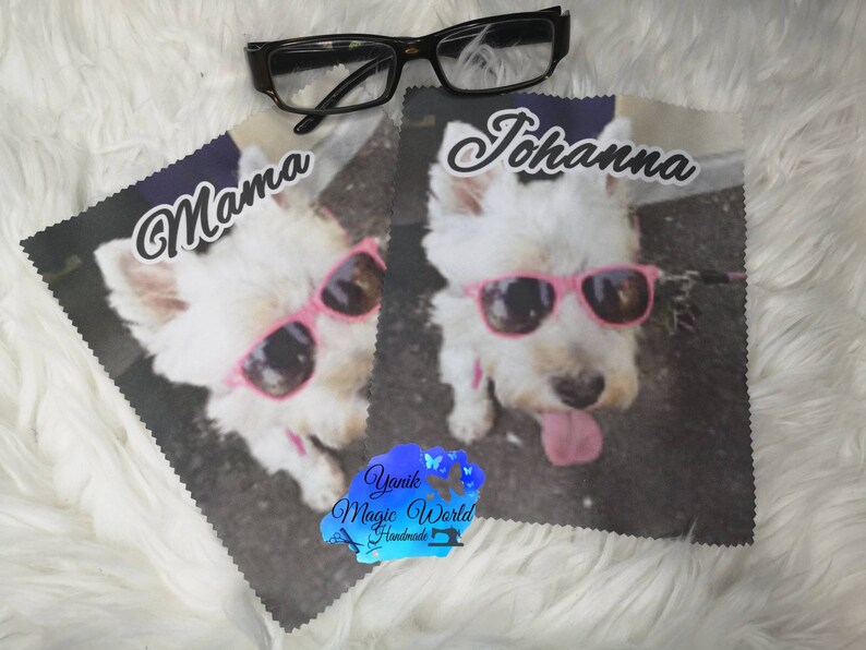 May include: Two microfiber cleaning cloths featuring a white dog wearing pink sunglasses. One cloth has the word "Mama" and the other has the name "Johanna". Black eyeglasses rest on top of the cloths. The cloths are on a white, fluffy surface.