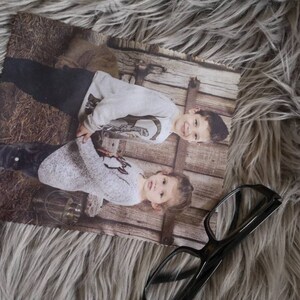 May include: A rectangular photo print featuring two children in light-colored sweaters and dark pants, set against a rustic wooden backdrop. The print is placed on a fluffy, gray surface, with a pair of eyeglasses resting nearby.