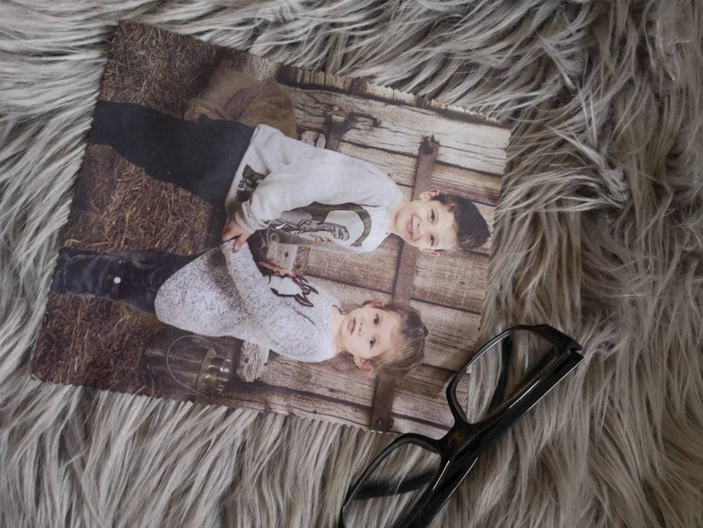 May include: A rectangular photo print featuring two children in light-colored sweaters and dark pants, set against a rustic wooden backdrop. The print is placed on a fluffy, gray surface, with a pair of eyeglasses resting nearby.