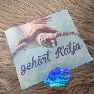 May include: A light blue square cloth featuring a sea turtle swimming underwater. The text "geh&ouml;rt Katja" is written in a dark blue script. A logo with the text "Yanik Magic World Handmade" is in the bottom right corner.