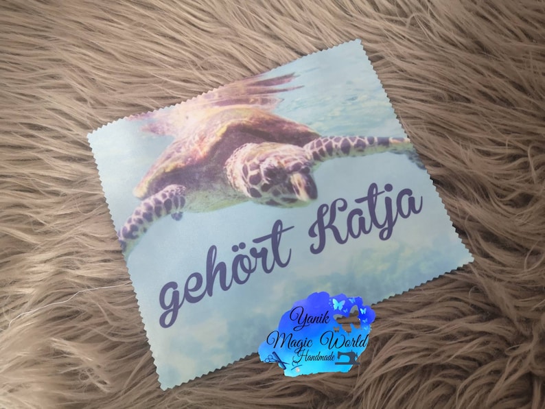 May include: A light blue square cloth featuring a sea turtle swimming underwater. The text "geh&ouml;rt Katja" is written in a dark blue script. A logo with the text "Yanik Magic World Handmade" is in the bottom right corner.
