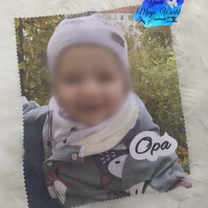 May include: A printed photo of a baby wearing a white hat, a scarf, and a gray jacket with a fox design. The photo has a white label with the word "Opa" on it. The photo is on a white, fluffy surface.